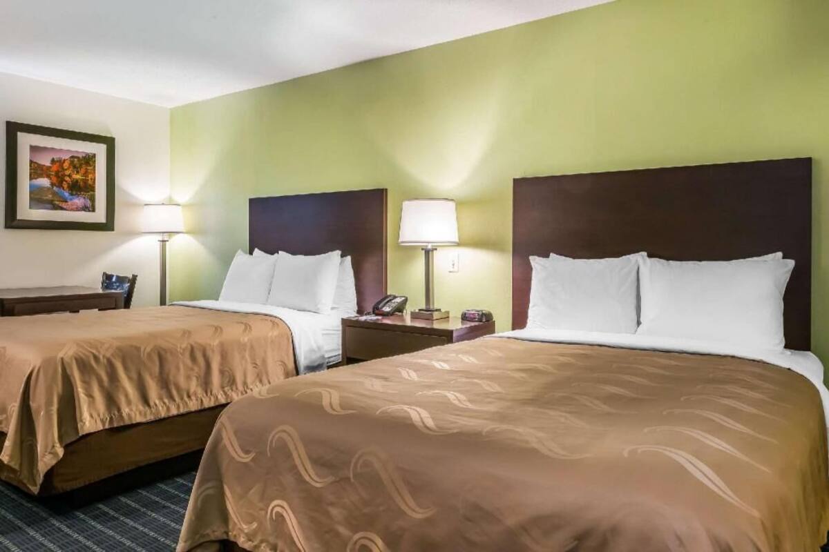 A hotel room with two beds, a desk, a chair, and a nightstand with a lamp.