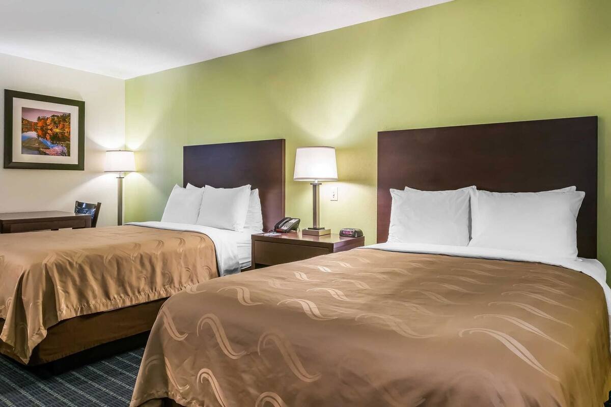 A hotel room with two beds, a desk, a chair, and a nightstand with a lamp.