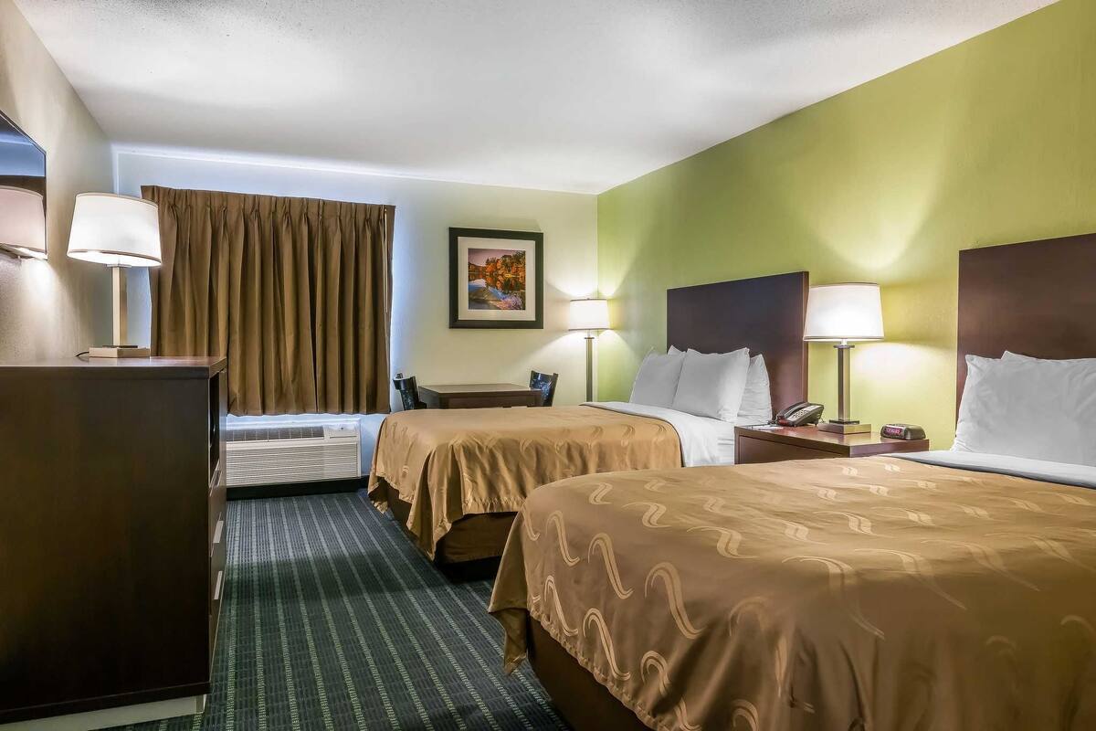 A hotel room with two beds, a desk, a chair, a lamp, a phone, and a picture on the wall.