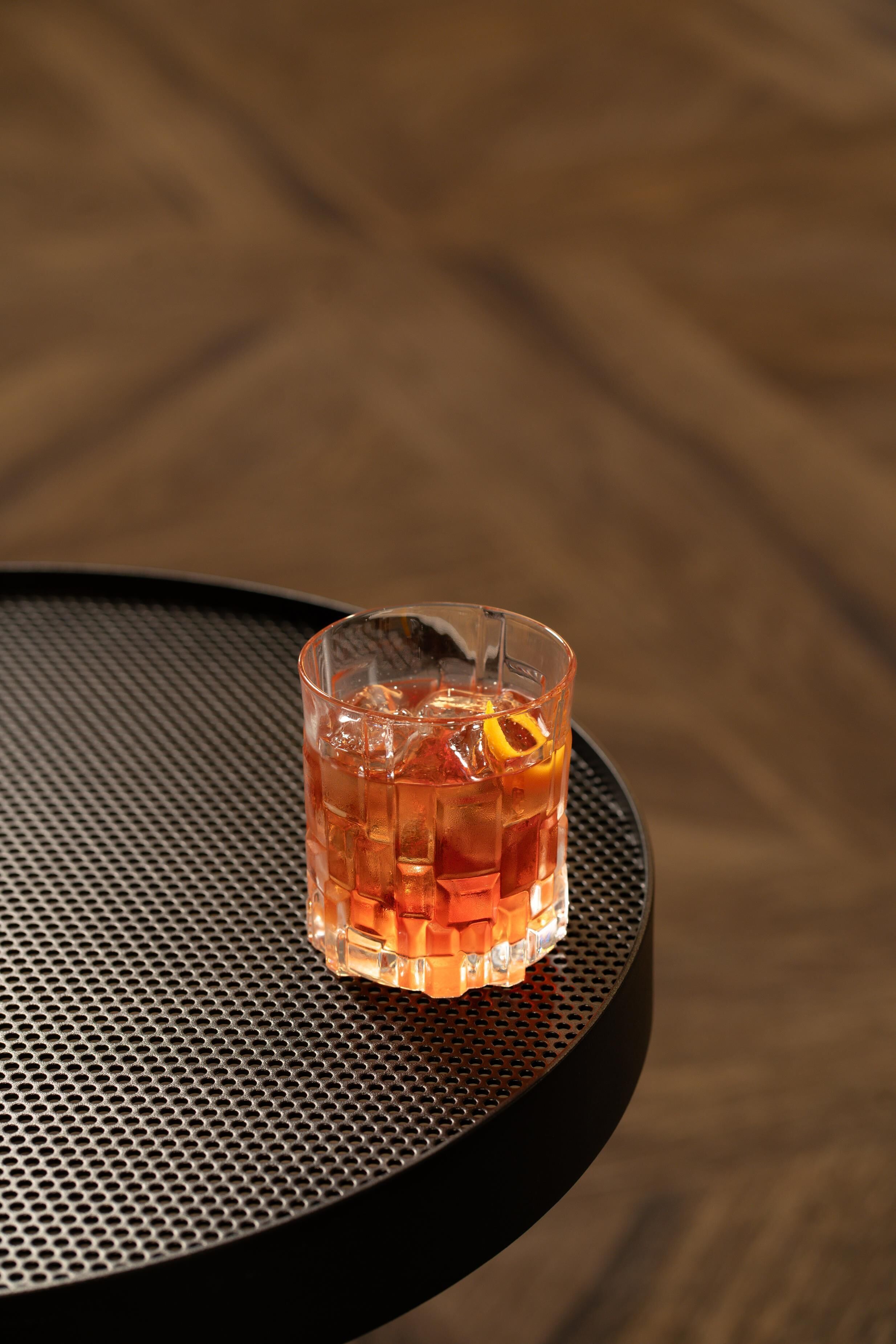 Un verre d’une boisson ambrée, avec des glaçons et une tranche d’orange, posé sur une table ronde en métal perforé.