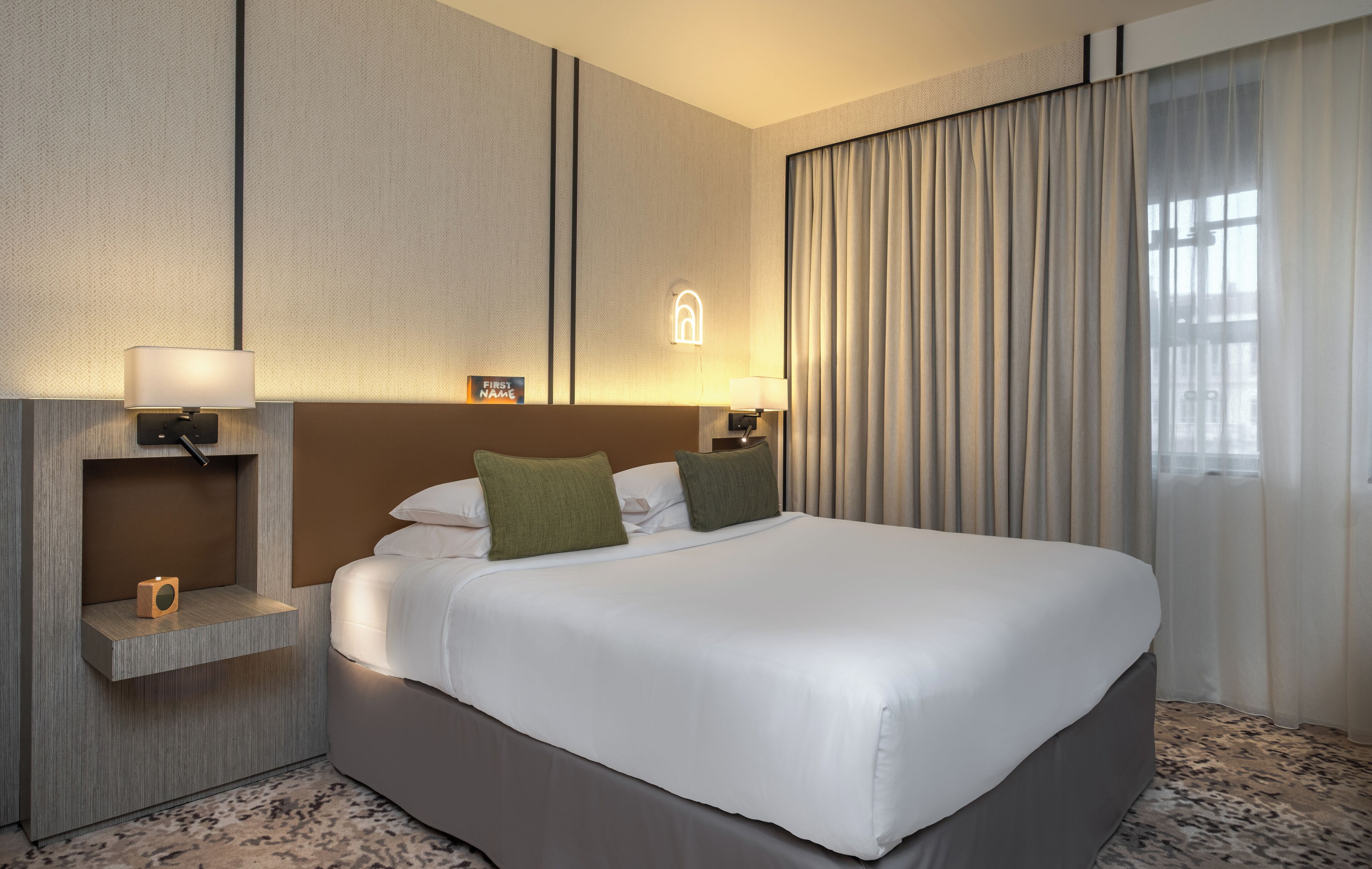 A hotel room with a large bed, two bedside tables with lamps, a small shelf with a lamp, and a window with curtains.