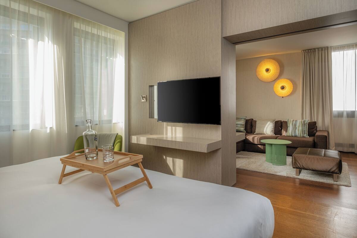 A modern hotel room with a large bed, a flat-screen TV mounted on the wall, a wooden side table with glasses and a bottle, a green side table, a brown leather ottoman, and a sofa with cushions.
