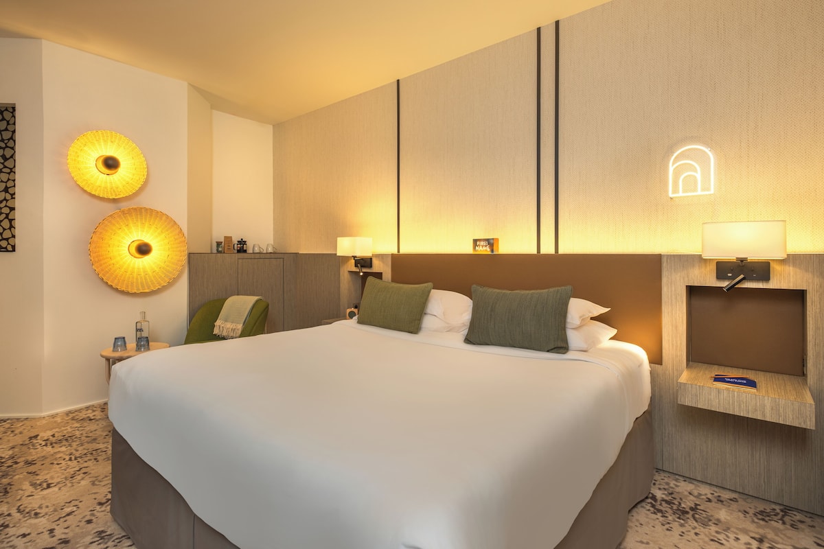 A hotel room with a large bed, two wall sconces, a nightstand, and a small shelf with a book.