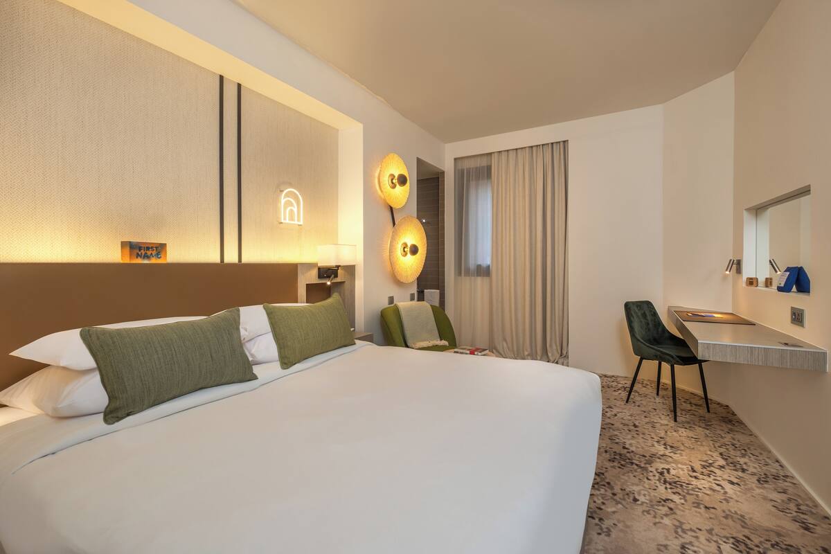 A modern hotel room with a large bed, a desk, and a chair. There is a mirror above the desk and a wall-mounted lamp.