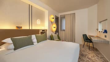 A modern hotel room with a large bed, a desk, and a chair. There is a mirror above the desk and a wall-mounted lamp.