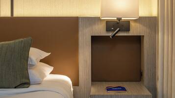 Habitación de hotel con una cama bien hecha, una lámpara de noche, un pequeño estante de madera con un libro y una lámpara de pared.