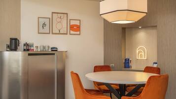 A modern hotel room with a round dining table, orange upholstered chairs, a stainless steel kitchenette, and artwork on the walls.