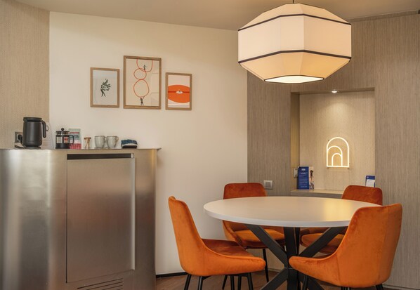 A modern hotel room with a round dining table, orange upholstered chairs, a stainless steel kitchenette, and artwork on the walls.