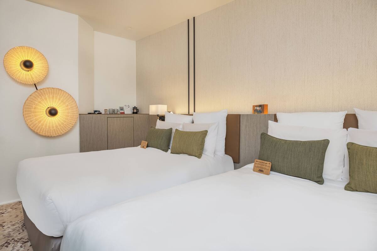 A hotel room with two beds, a nightstand, a lamp, and two wall-mounted lamps.