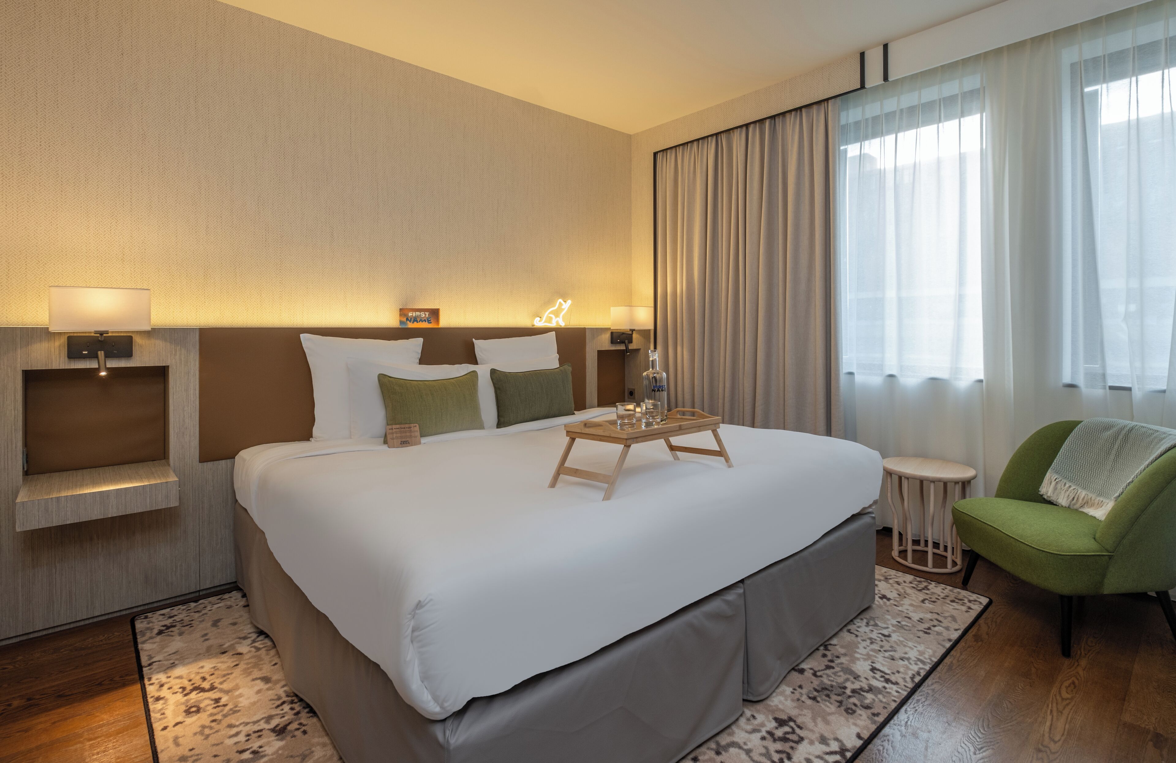 Une chambre d’hôtel avec un grand lit, deux tables de chevet avec des lampes, un fauteuil vert et une fenêtre avec des rideaux.