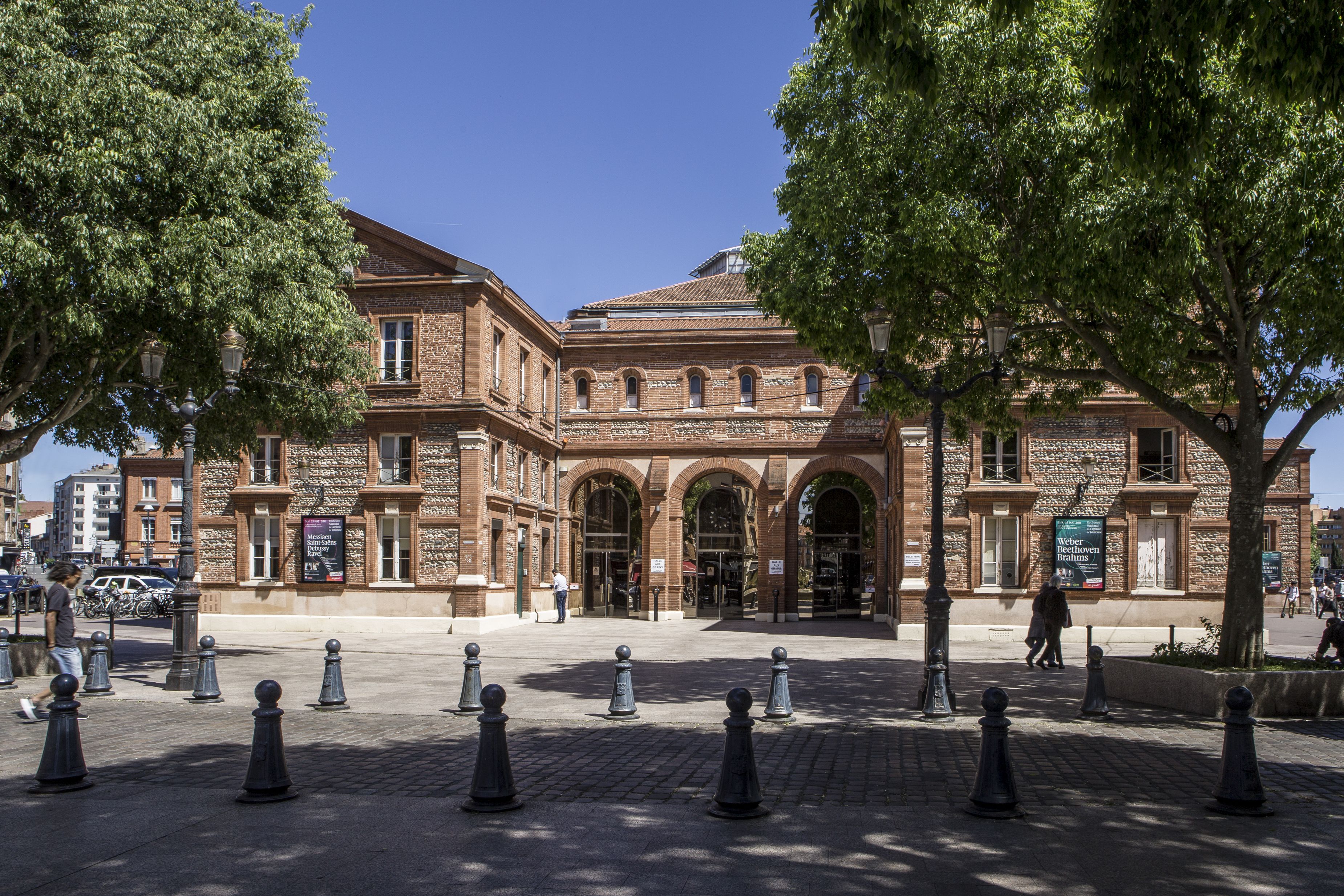 A historic building with arched entrances, a brick facade, and a paved courtyard with bollards.