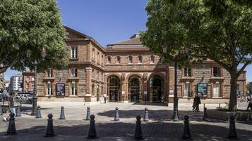 A historic building with arched entrances, a brick facade, and a paved courtyard with bollards.