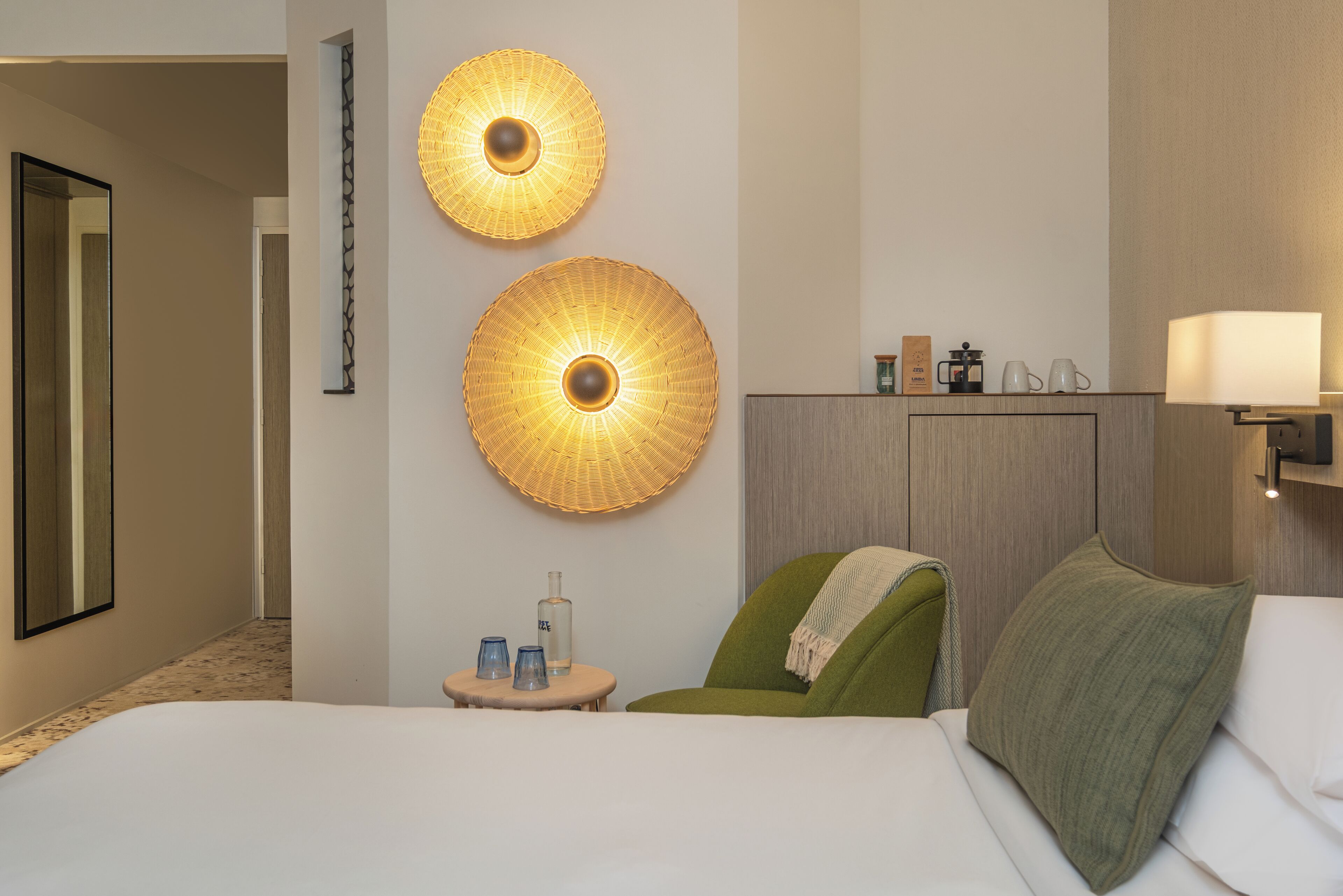 Een hotelkamer met een bed, een groene fauteuil, een ronde wandlamp, een nachtkastje met een fles en glazen, en een nachtkastje met een lamp.