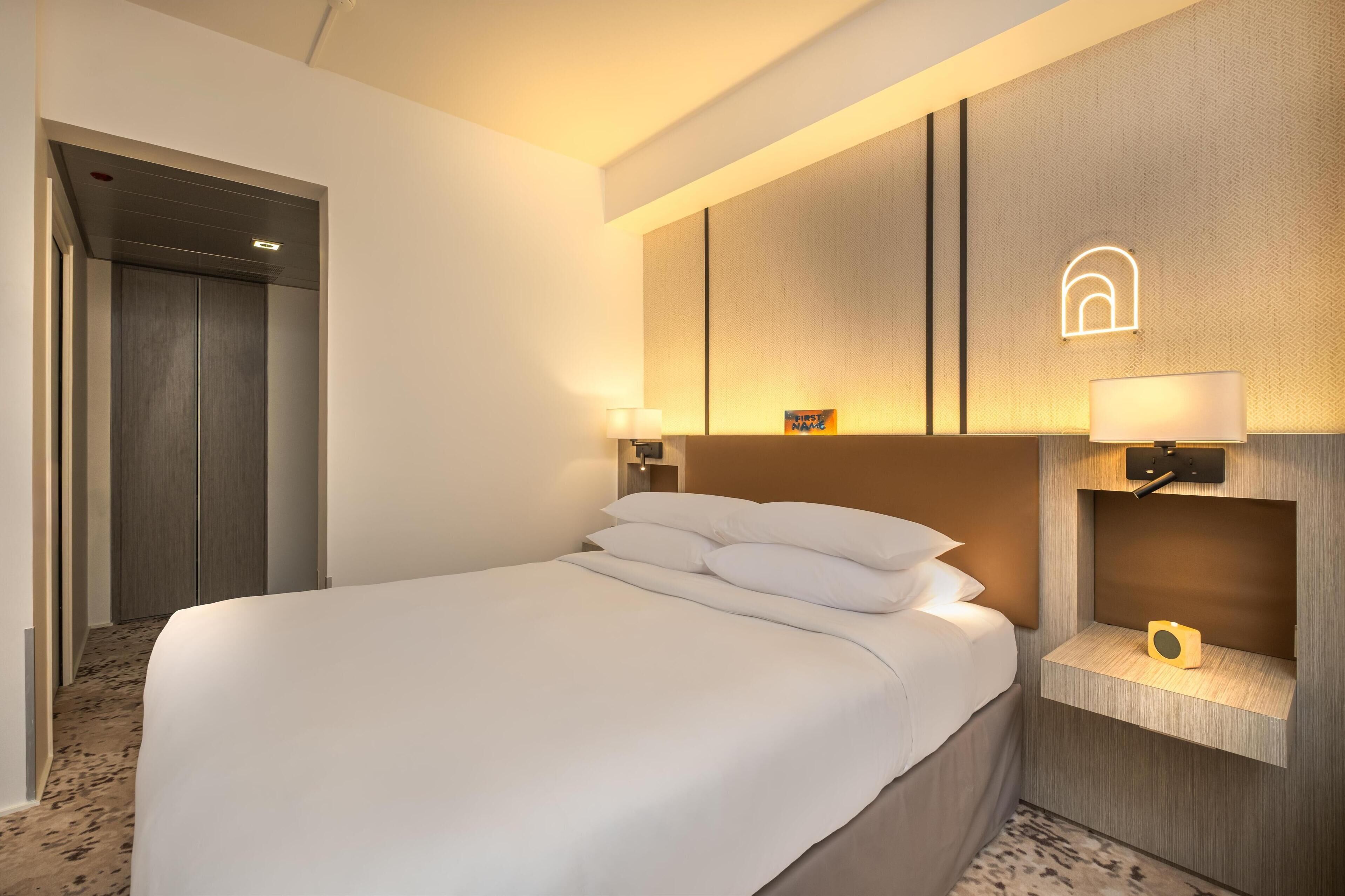Habitación de hotel con una cama grande, una mesita de noche con lámpara, un estante en la pared y una puerta que da a otra habitación.