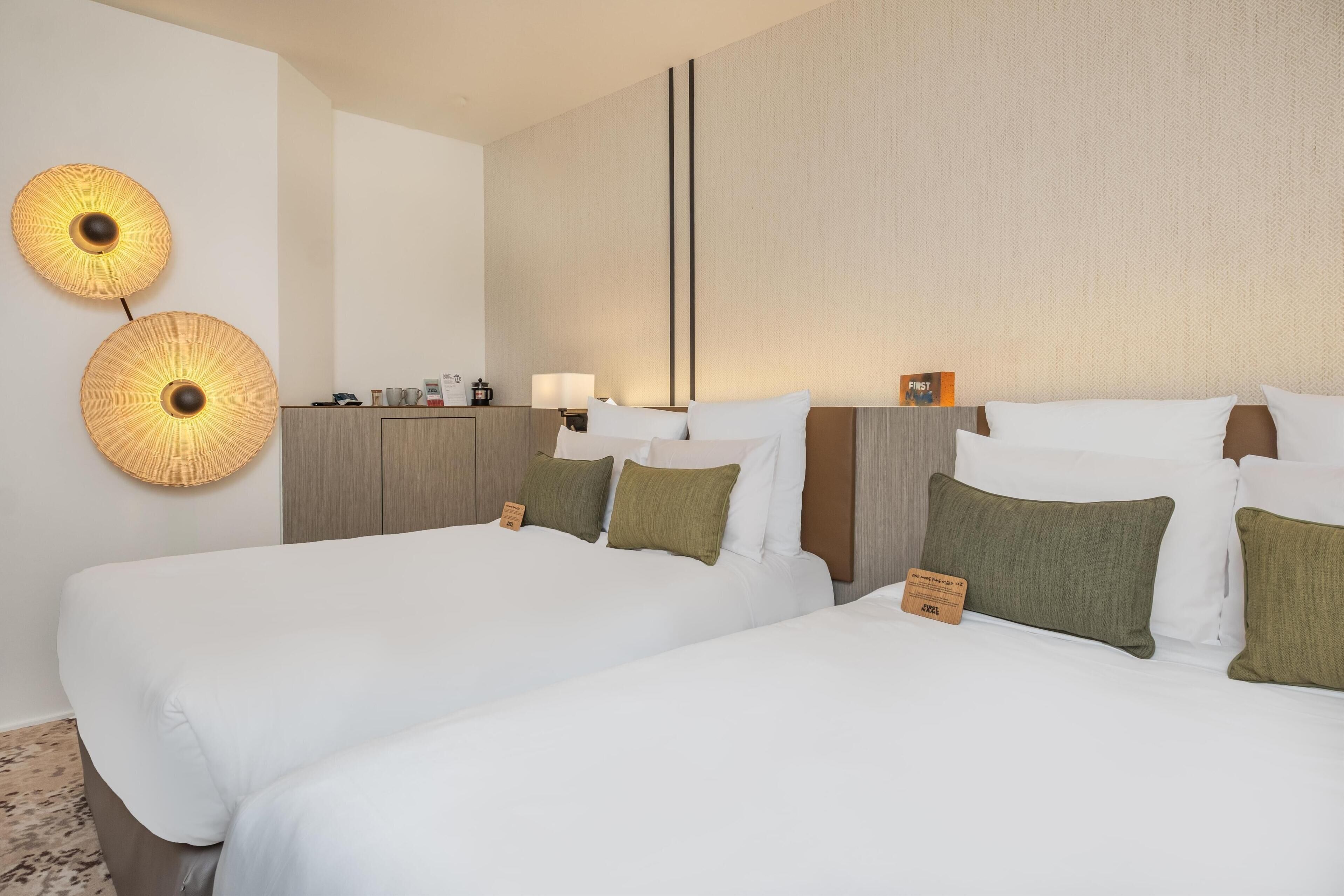 A hotel room with two beds, a nightstand, a lamp, and two wall-mounted lamps.