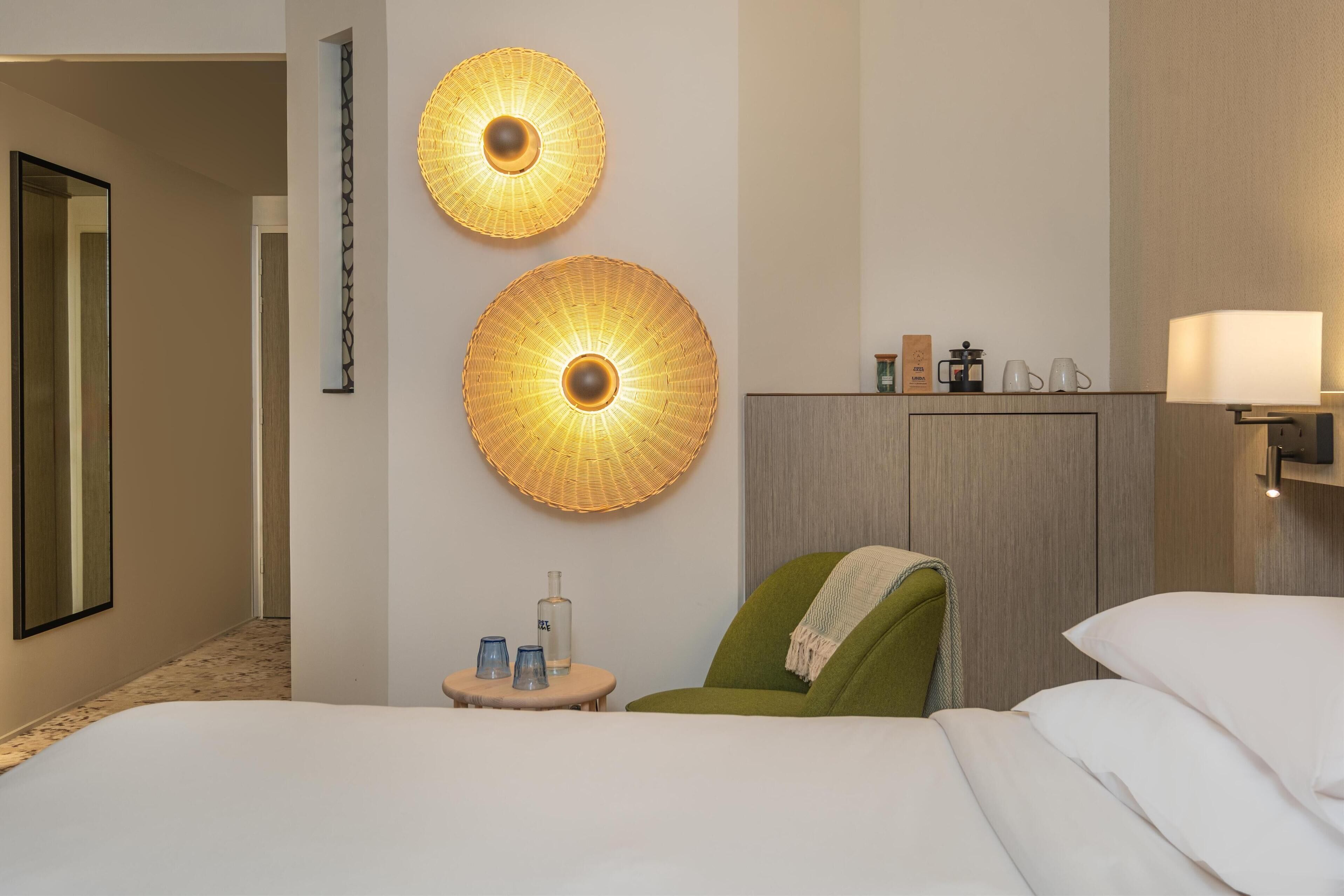 A hotel room with a bed, a green armchair, two wall-mounted lamps, a small table with a bottle and glasses, and a minibar.