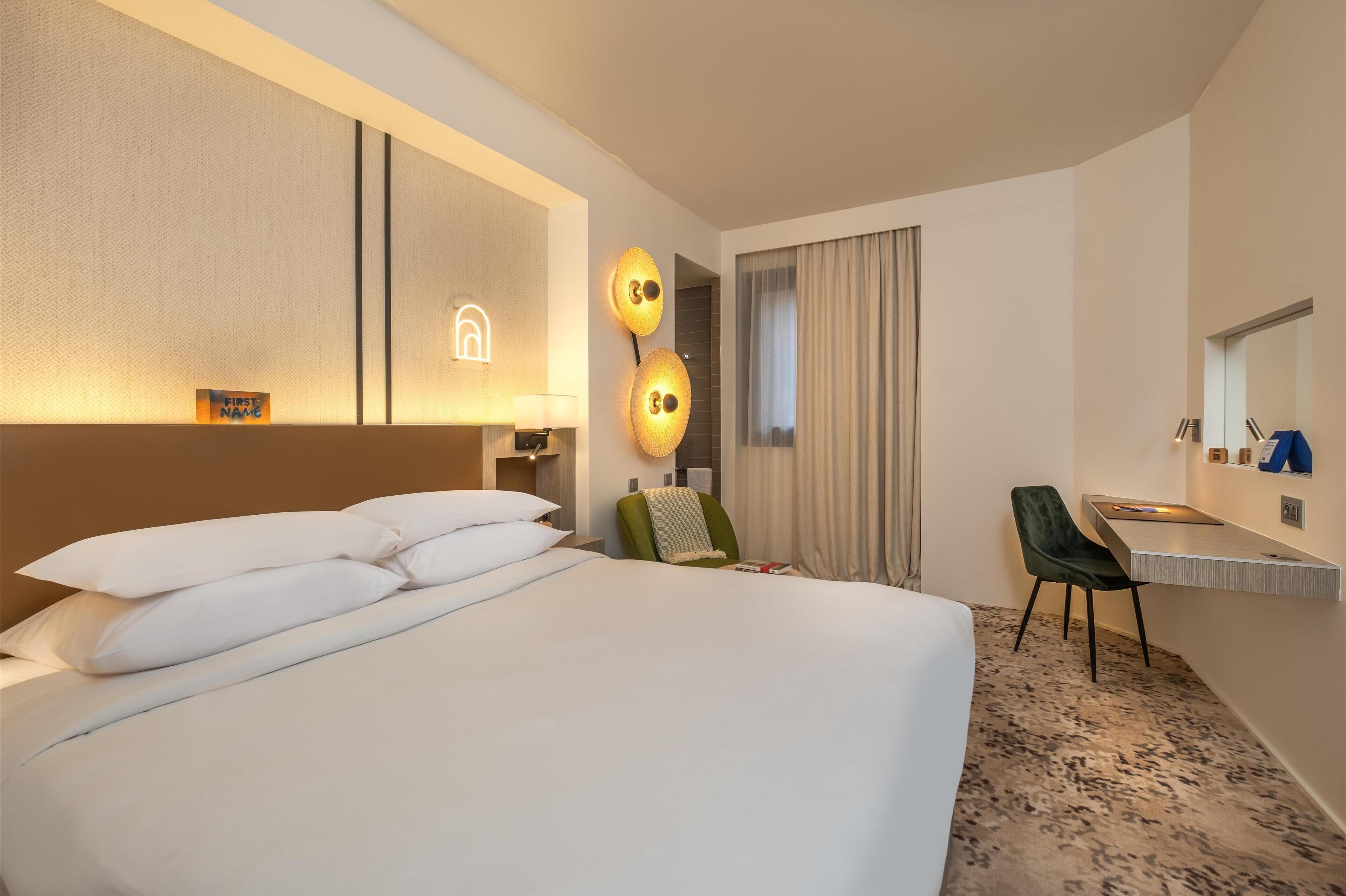 A hotel room with a large bed, a desk, a chair, a mirror, and a wall-mounted light fixture.