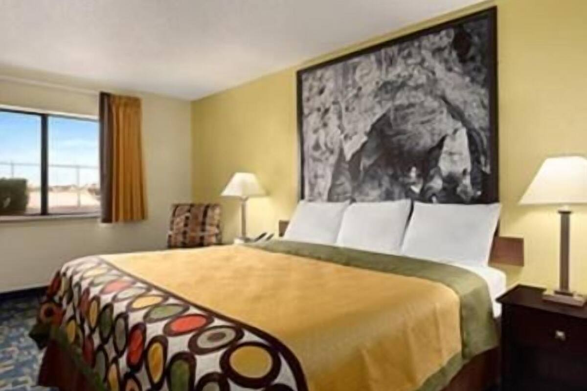 A hotel room with a bed, a window with curtains, a painting on the wall, and a lamp on a bedside table.