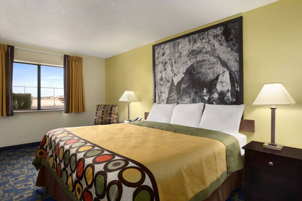 A hotel room with a large bed, two bedside lamps, a large painting on the wall, and a window with curtains.