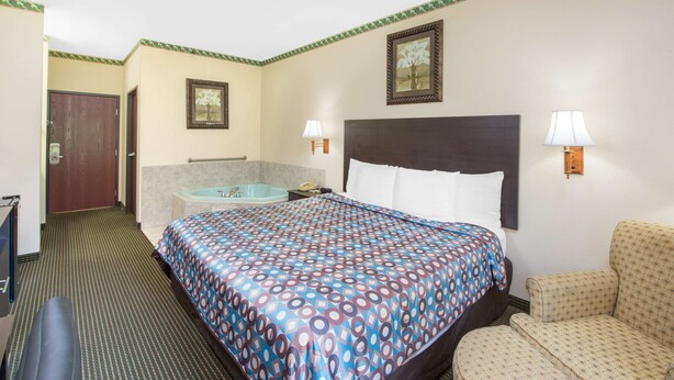 A hotel room with a large bed, a chair, a desk, a desk lamp, a nightstand, a framed picture, and a wall-mounted lamp.