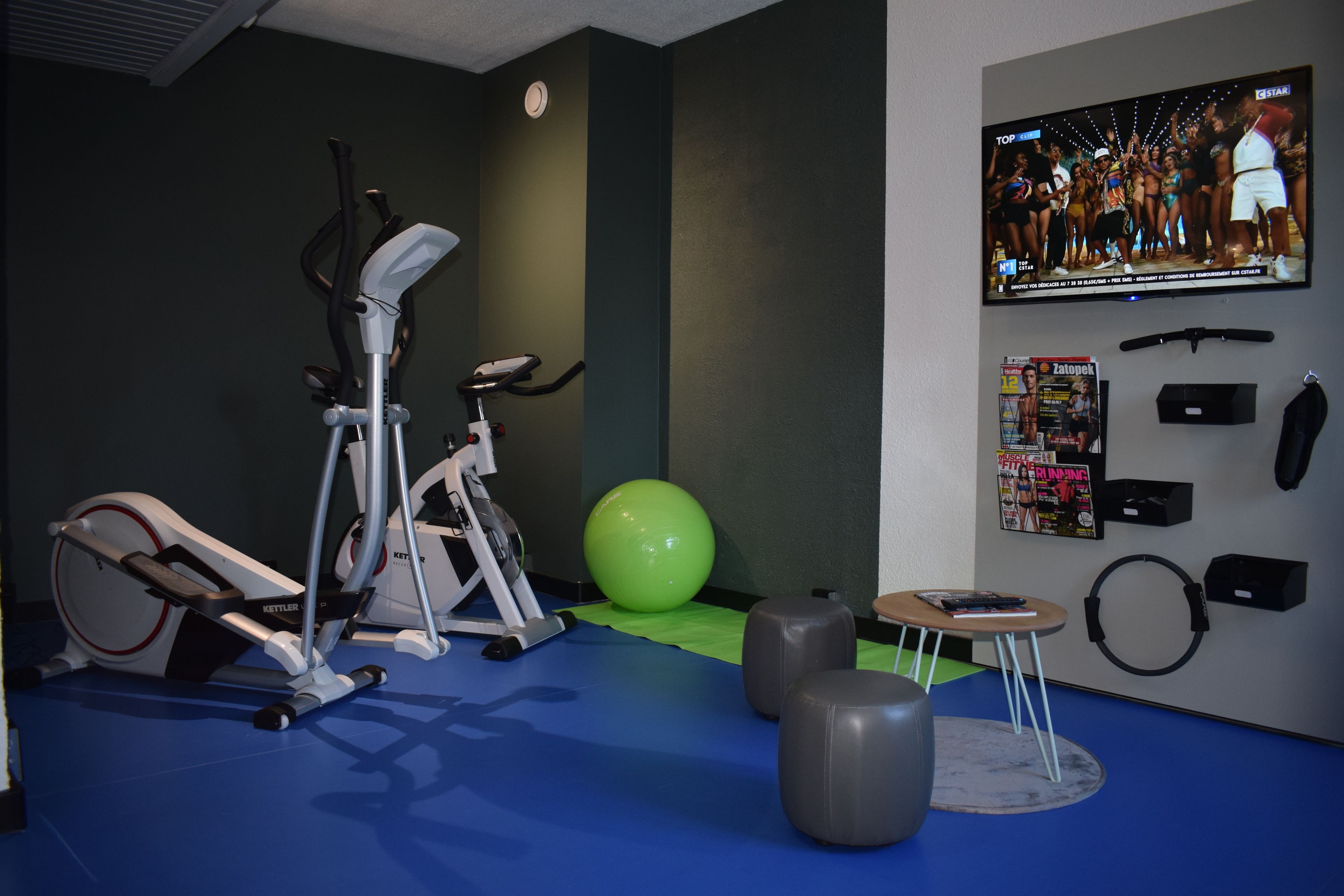 Ein Fitnessraum mit Trainingsgeräten, einem grünen Übungsball und einem Fernseher an der Wand.