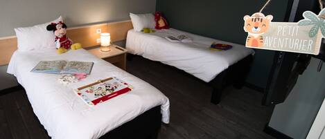 A hotel room with two beds, each with a white bedspread and pillows. There is a Mickey Mouse plush toy on the left bed, and a sign with a tiger illustration on the right bed. A lamp is on the left nightstand, and a book is on the right bed.