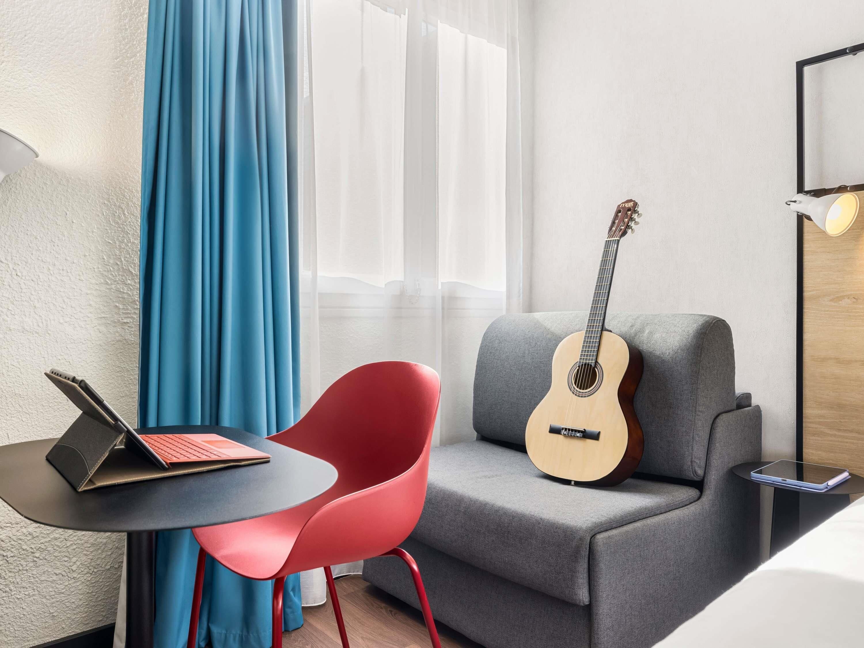 Una stanza moderna con un divano grigio, una poltrona rossa, un laptop su un tavolino e una chitarra appoggiata al muro.