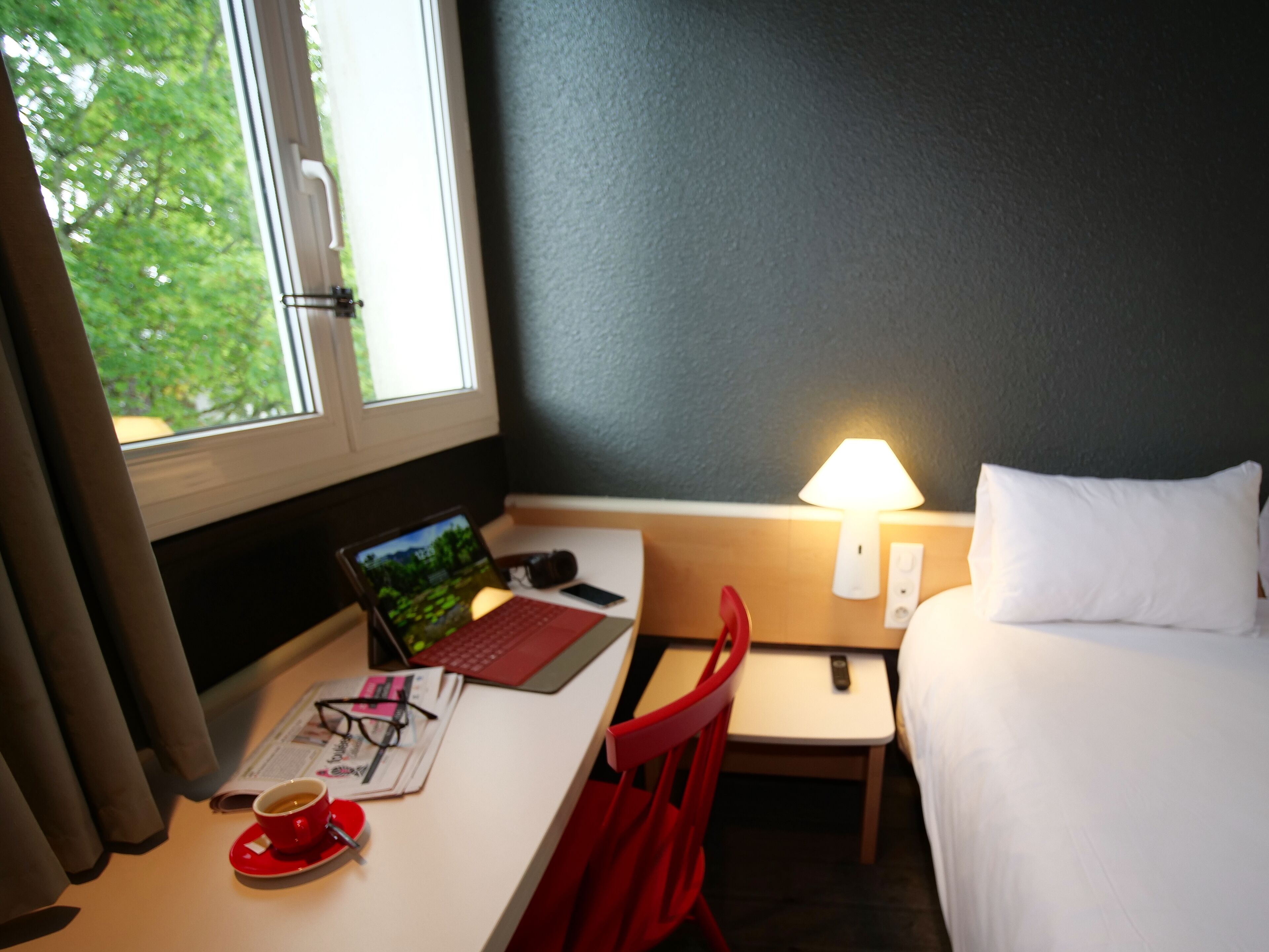 Een hotelkamer met een bureau, laptop, rode stoel, nachtkastje met een lamp, en een bed met witte kussens.