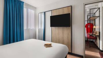 Habitación de hotel con cama, televisor montado en la pared, escritorio con silla y armario.