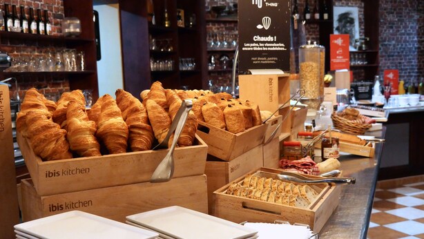 Mostrador de panadería con croissants y otros productos horneados, cajas de madera con la etiqueta 'ibis kitchen' y pilas de platos blancos.