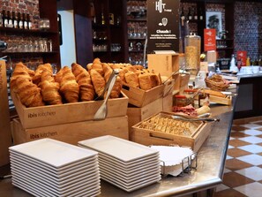 A bakery display with croissants and other baked goods, wooden boxes labeled 'ibis kitchen', and stacks of white plates.