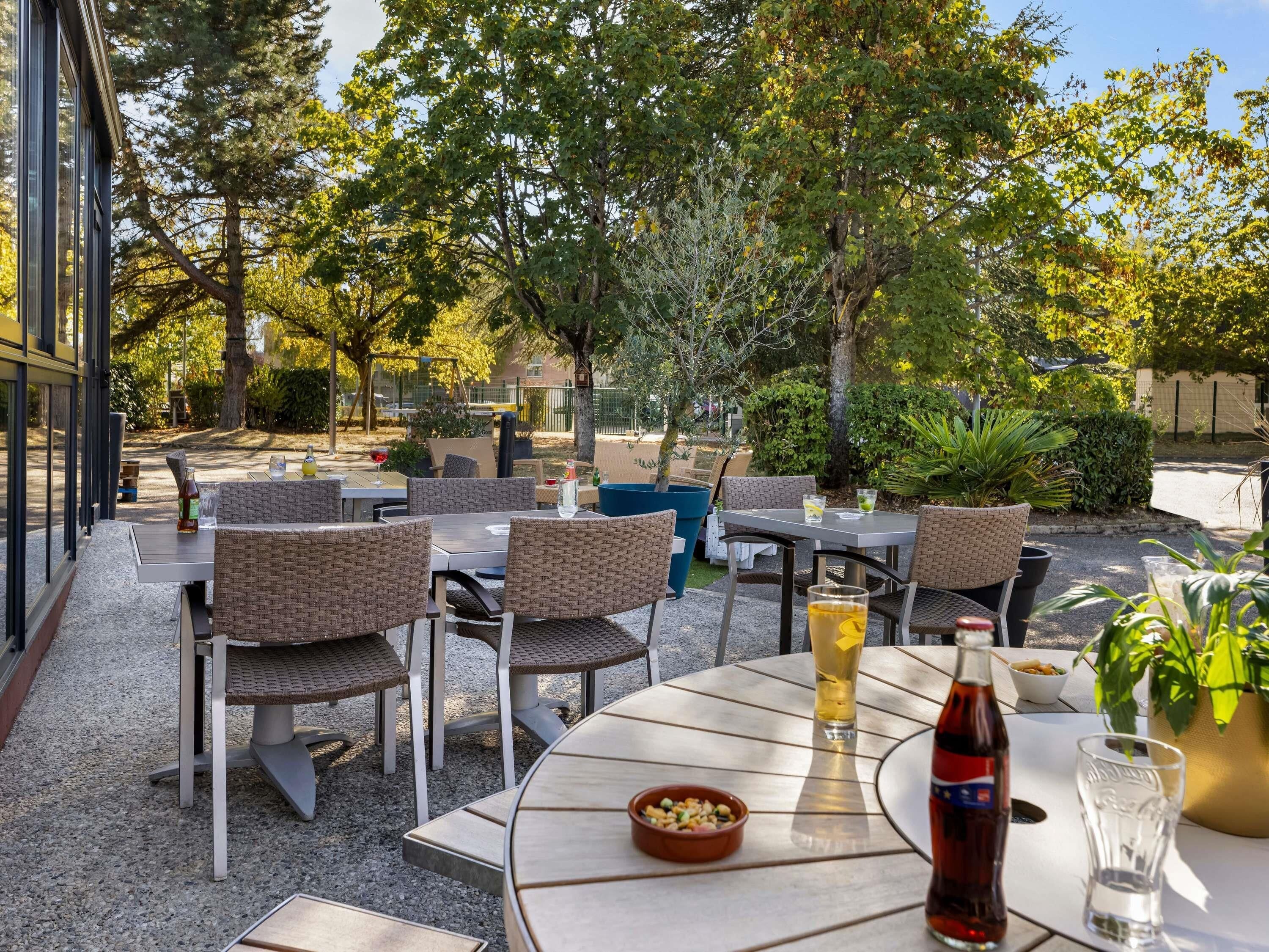 Een terras buiten met ronde tafels en rieten stoelen, omgeven door bomen en begroeiing.