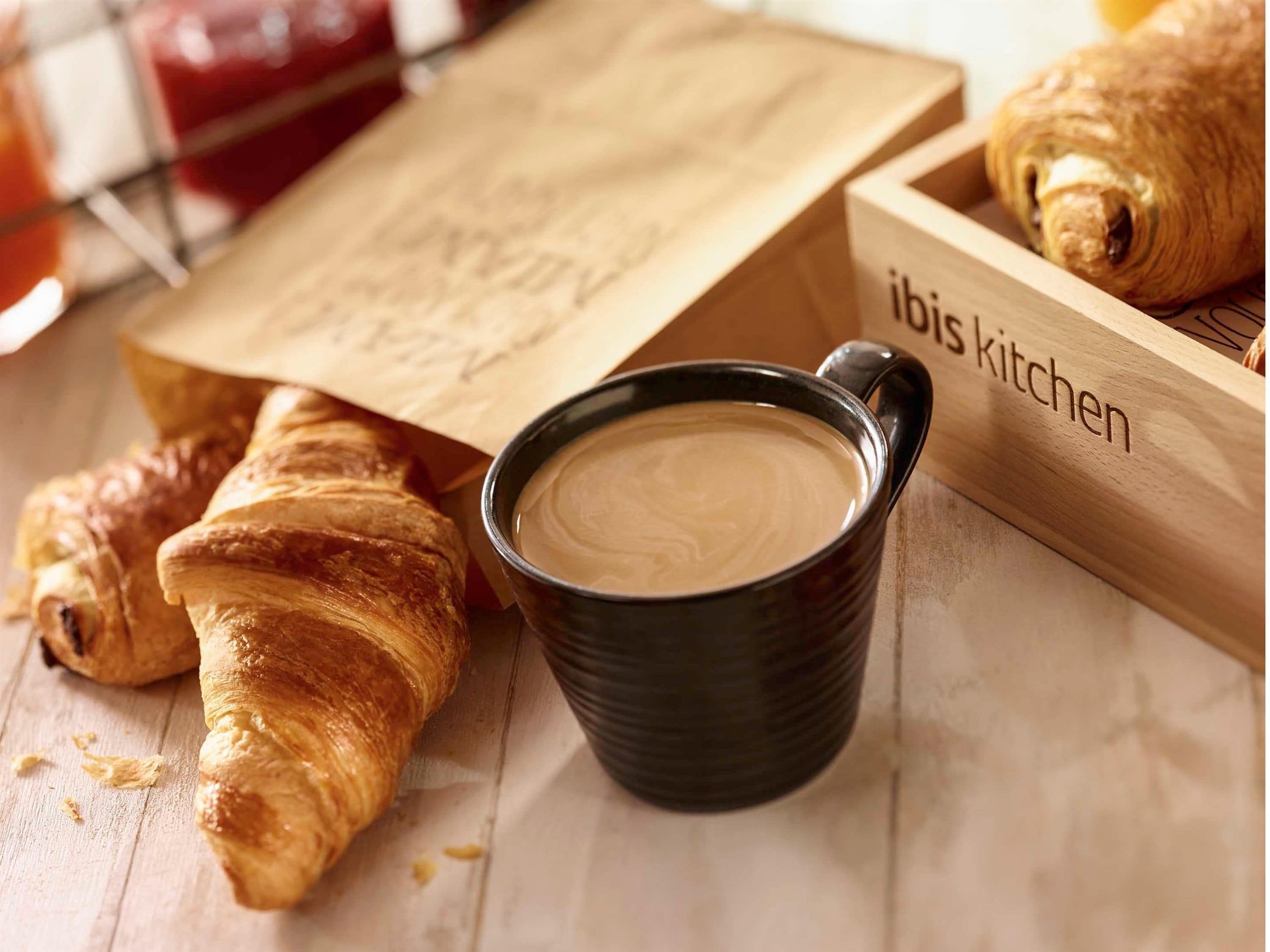 Une boîte en bois portant l’inscription « Ibis Kitchen », avec des croissants et une tasse de café posés sur une surface en bois.