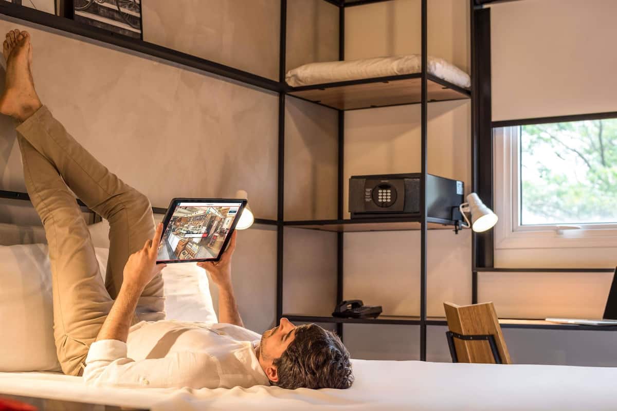 Uma pessoa deitada numa cama em quarto com janela, escrivaninha e prateleira com luminária.