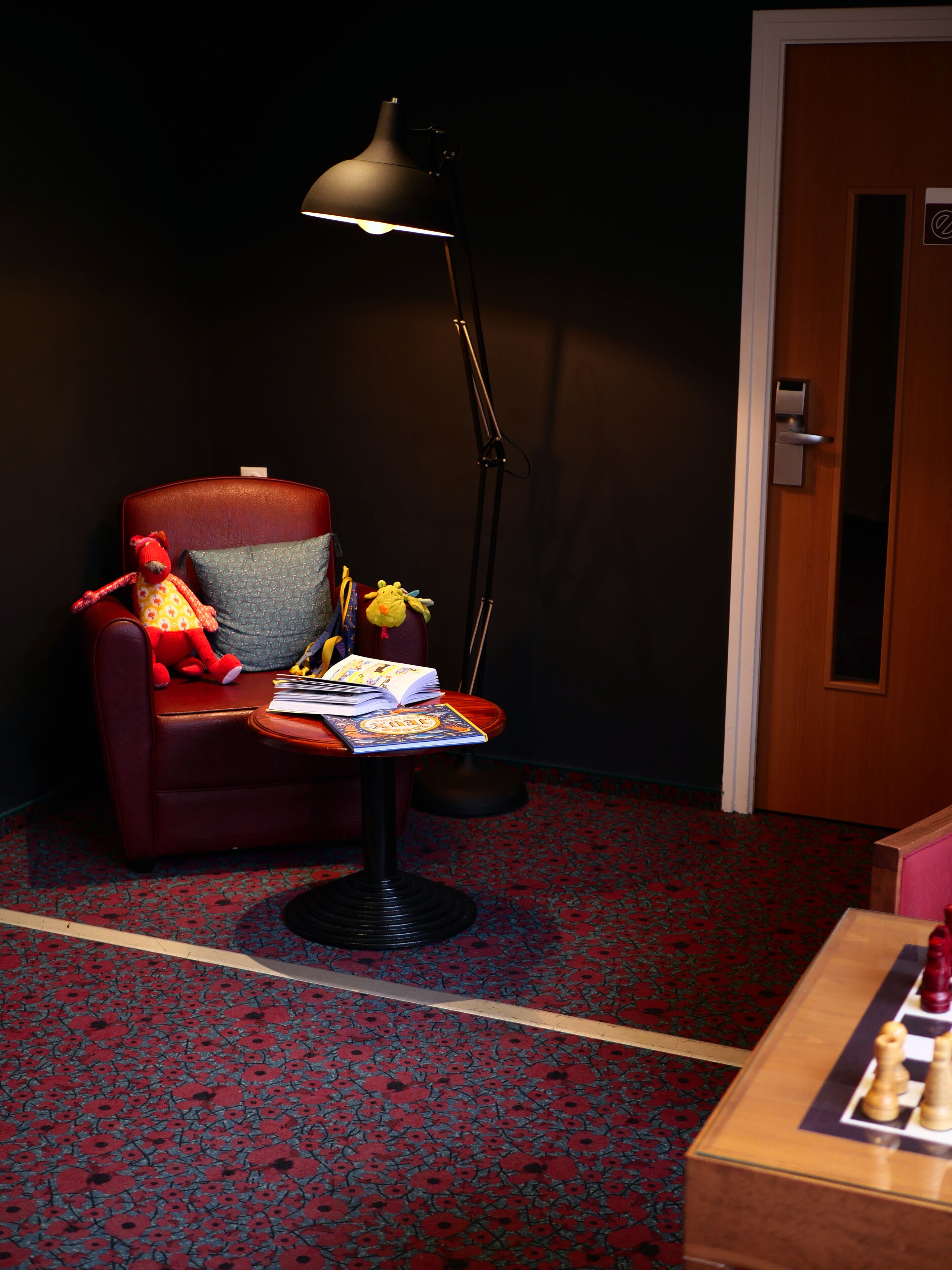 A room with a red armchair, a small table with books and a lamp, and a chessboard on a wooden table.