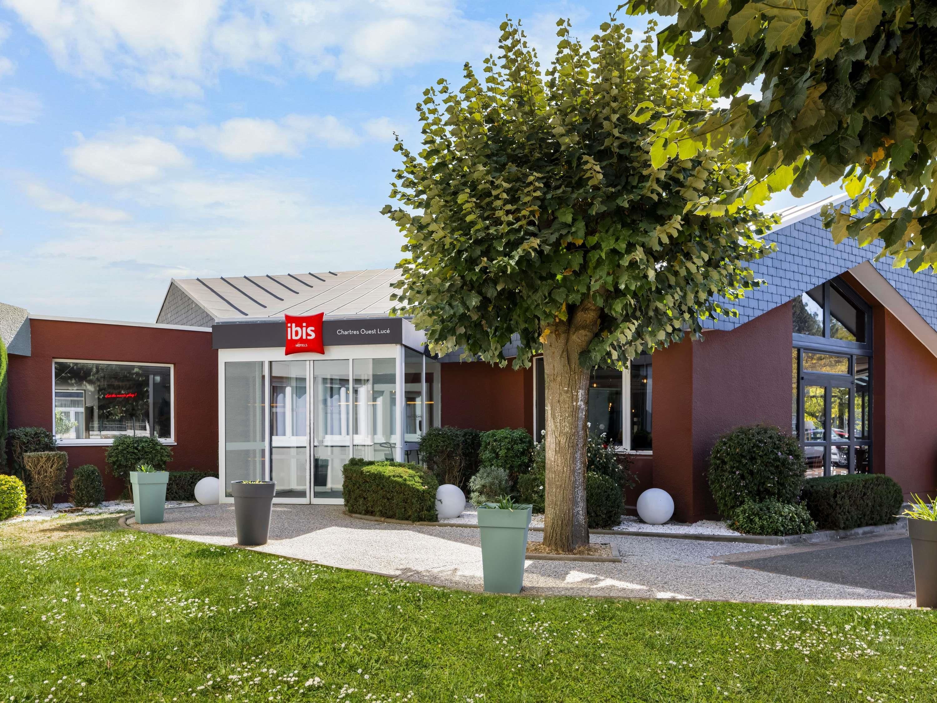 A hotel with a modern design, featuring a glass entrance, a red logo, and a well-maintained garden with a large tree and spherical planters.