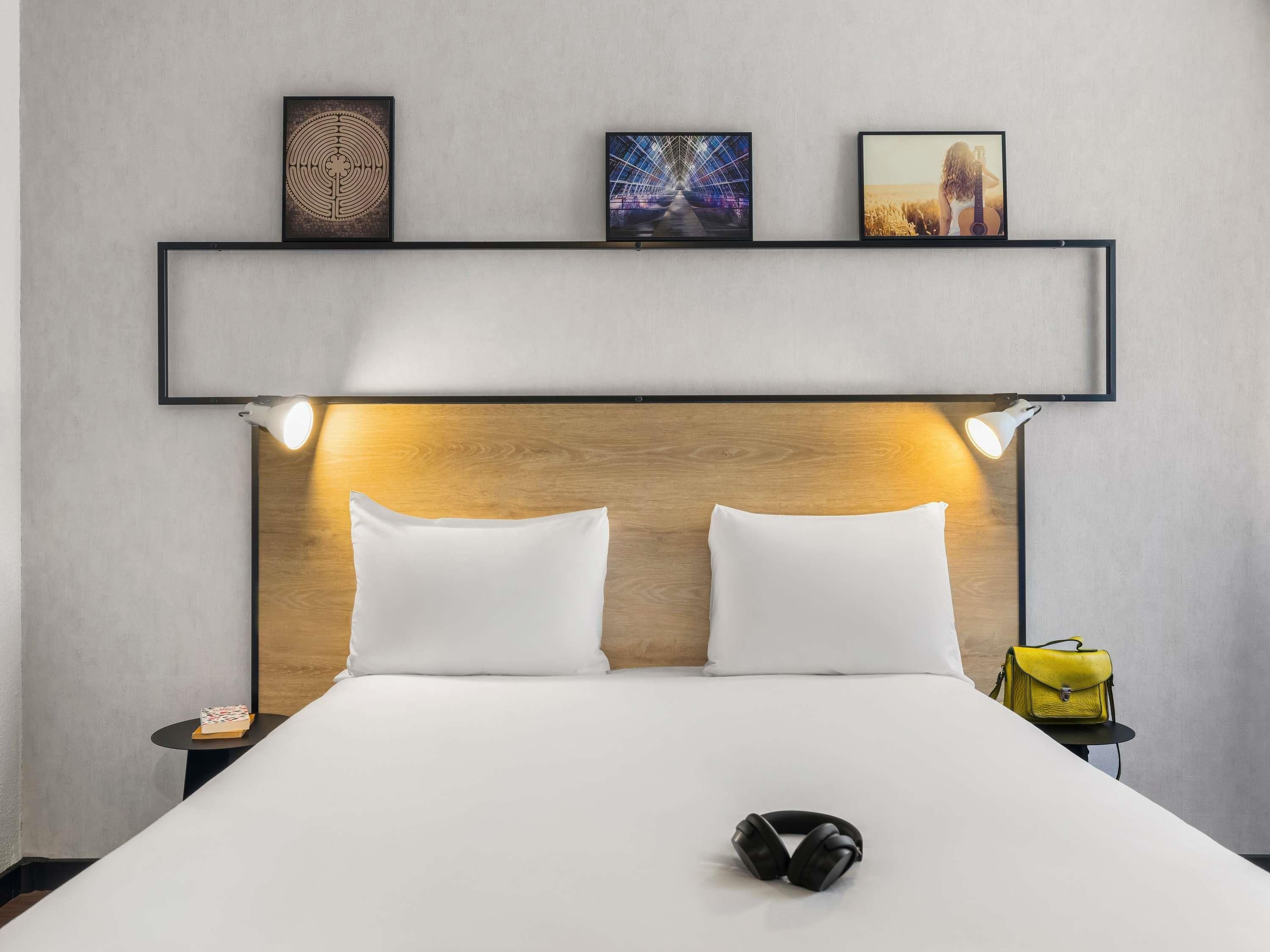 Une chambre d’hôtel comprenant un lit, deux lampes de chevet, une tête de lit et une étagère avec trois tableaux encadrés.