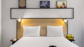 Une chambre d’hôtel comprenant un lit, deux lampes de chevet, une tête de lit et une étagère avec trois tableaux encadrés.