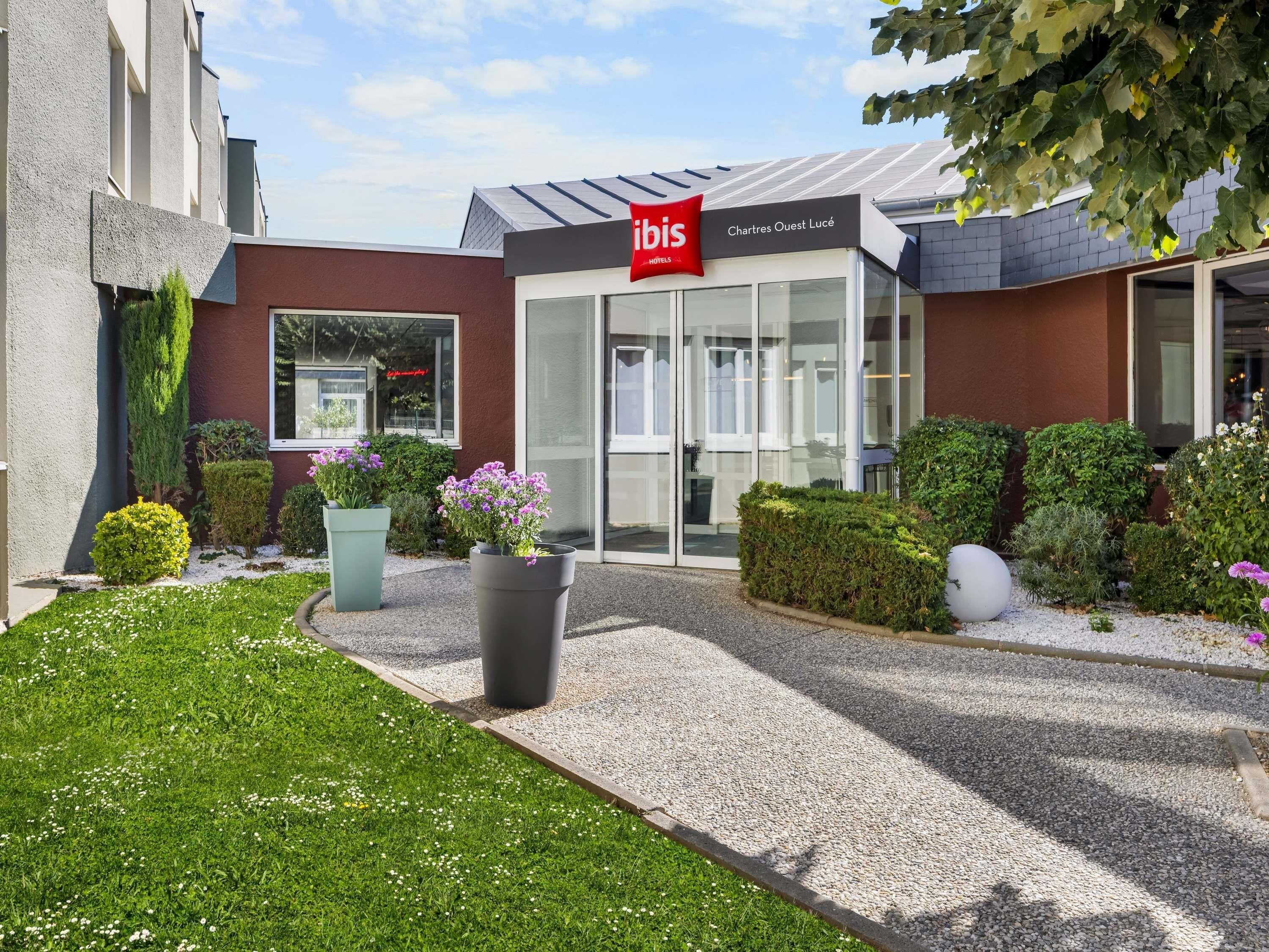 Een hotelentree met een modern ontwerp, een heldere glazen gevel en een verzorgde tuin met diverse planten en bloemen.