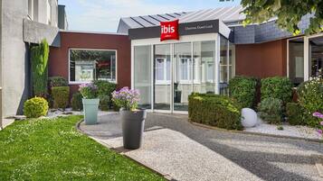 Een hotelentree met een modern ontwerp, een heldere glazen gevel en een verzorgde tuin met diverse planten en bloemen.