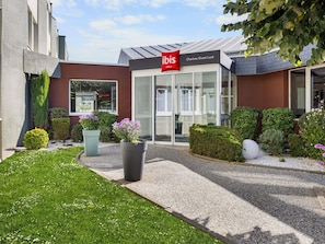 A hotel entrance with a modern design, a clear glass facade, and a well-maintained garden with various plants and flowers. - ibis Chartres Ouest Luce (Lucé)