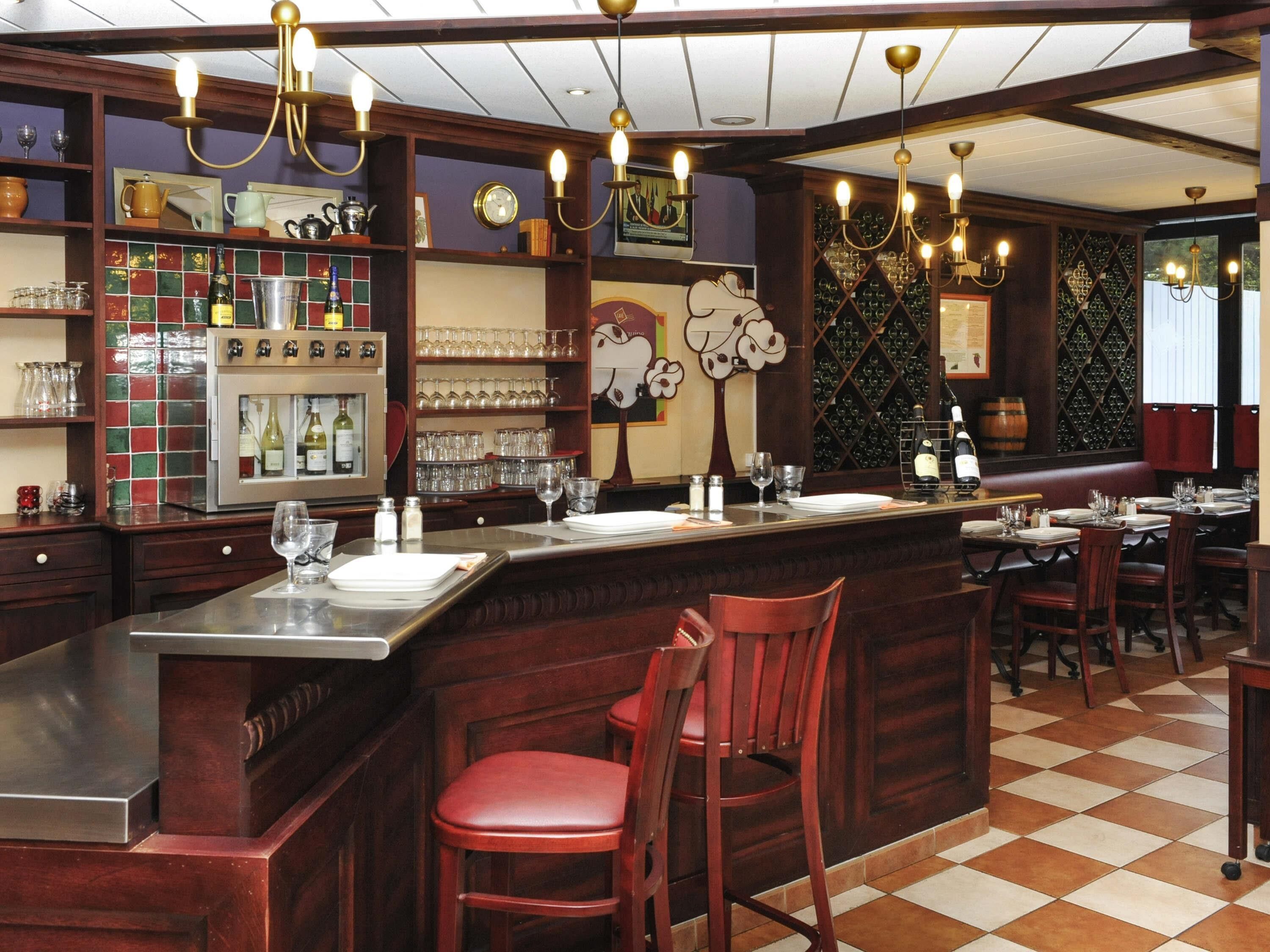 Uma área de bar com piso xadrez, balcão de madeira e banquetas vermelhas. Há taças de vinho e garrafas nas prateleiras, e um suporte de vinhos ao fundo.