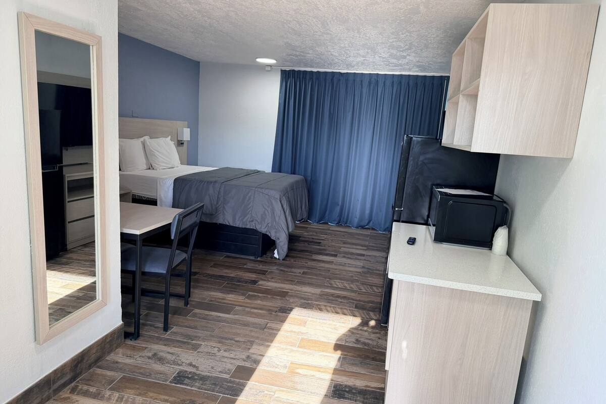 A compact hotel room with a bed, a desk, a chair, a small kitchenette, and a wardrobe.