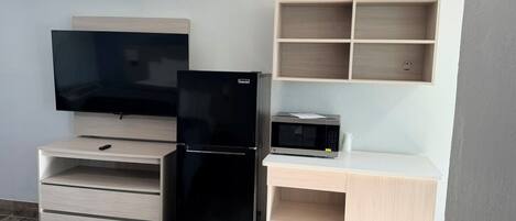 A compact room with a flat-screen TV, a black refrigerator, a microwave, and a wooden desk with open shelving.