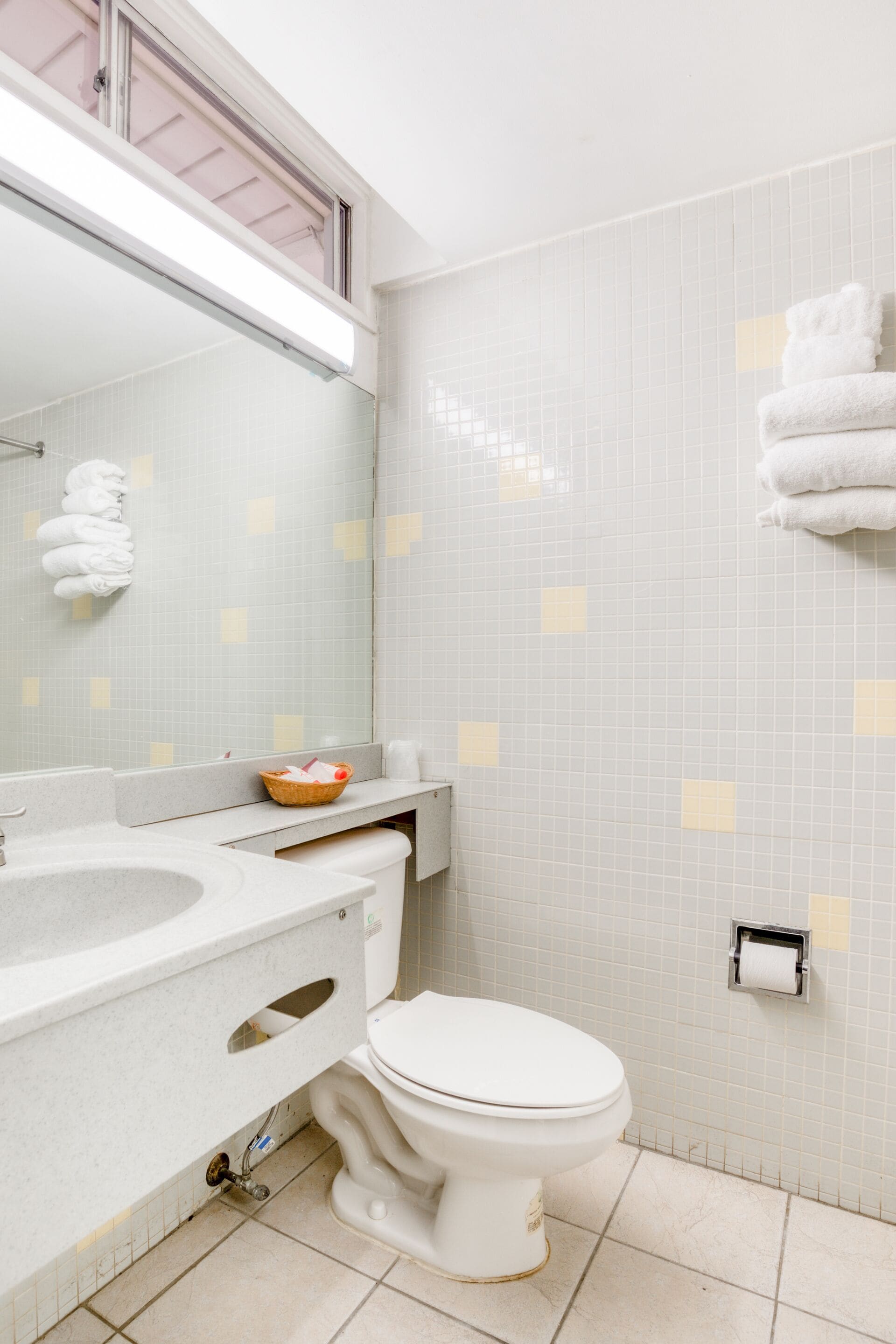 Een badkamer met een wit wastafel, toilet en een spiegel boven het wastafel. Handdoeken hangen aan de muur en er staat een mandje op het aanrecht.