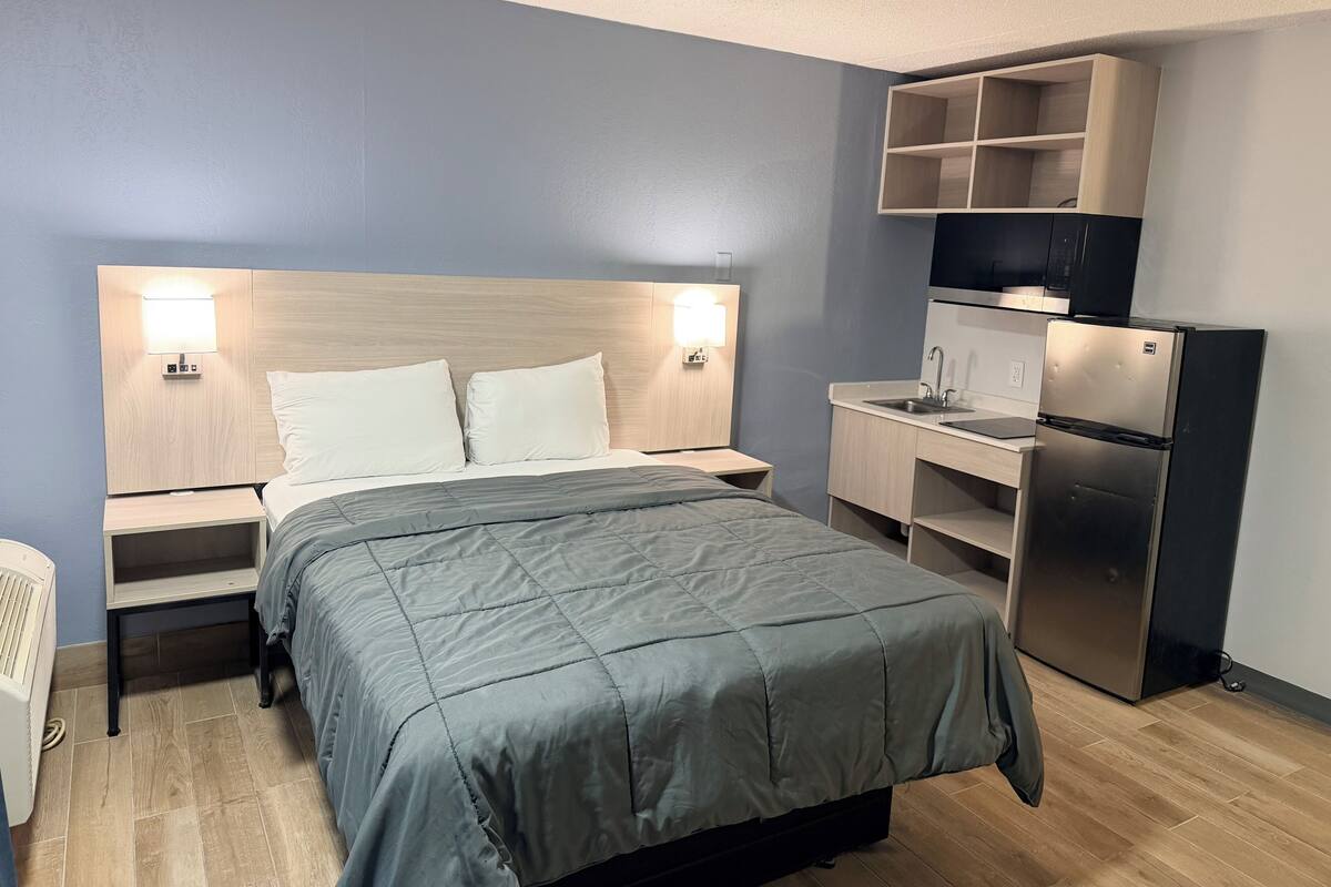 A hotel room with a bed, bedside tables, a desk, a refrigerator, and a built-in TV shelf.
