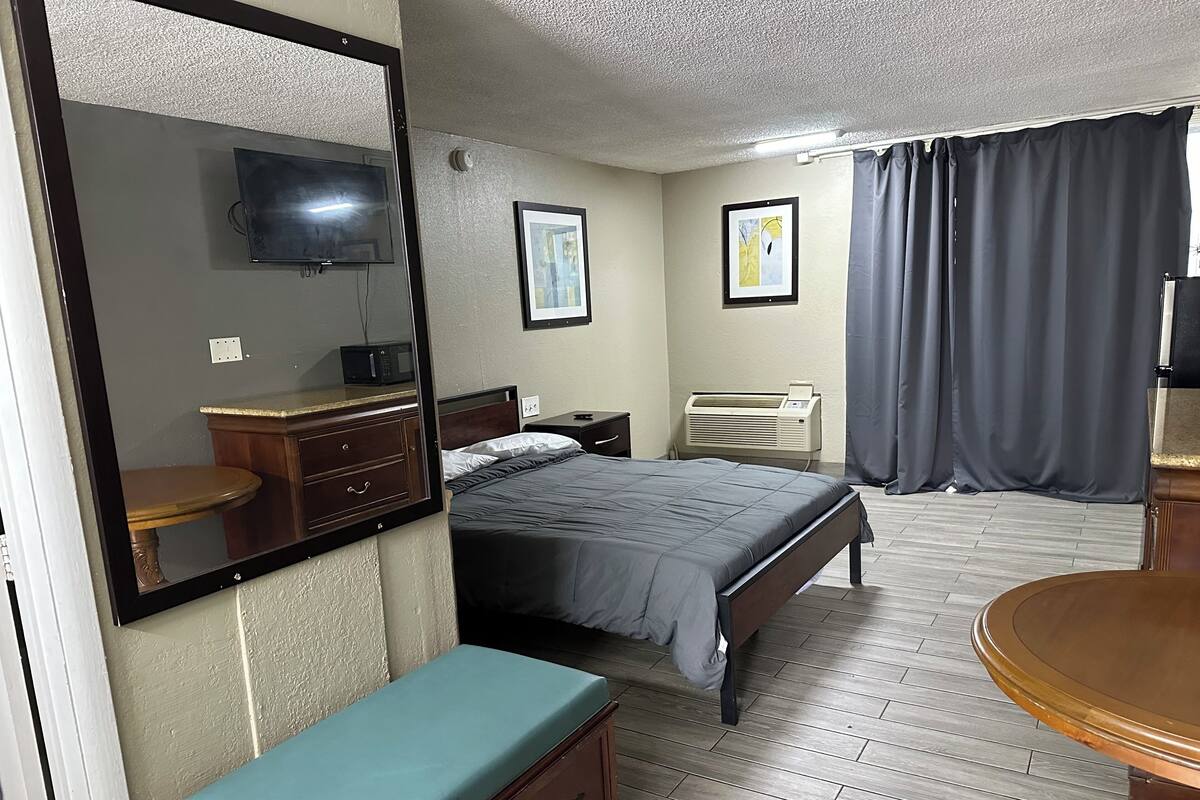 A bedroom with a bed, a bedside table, a dresser, a mirror, a television, and a window with curtains.