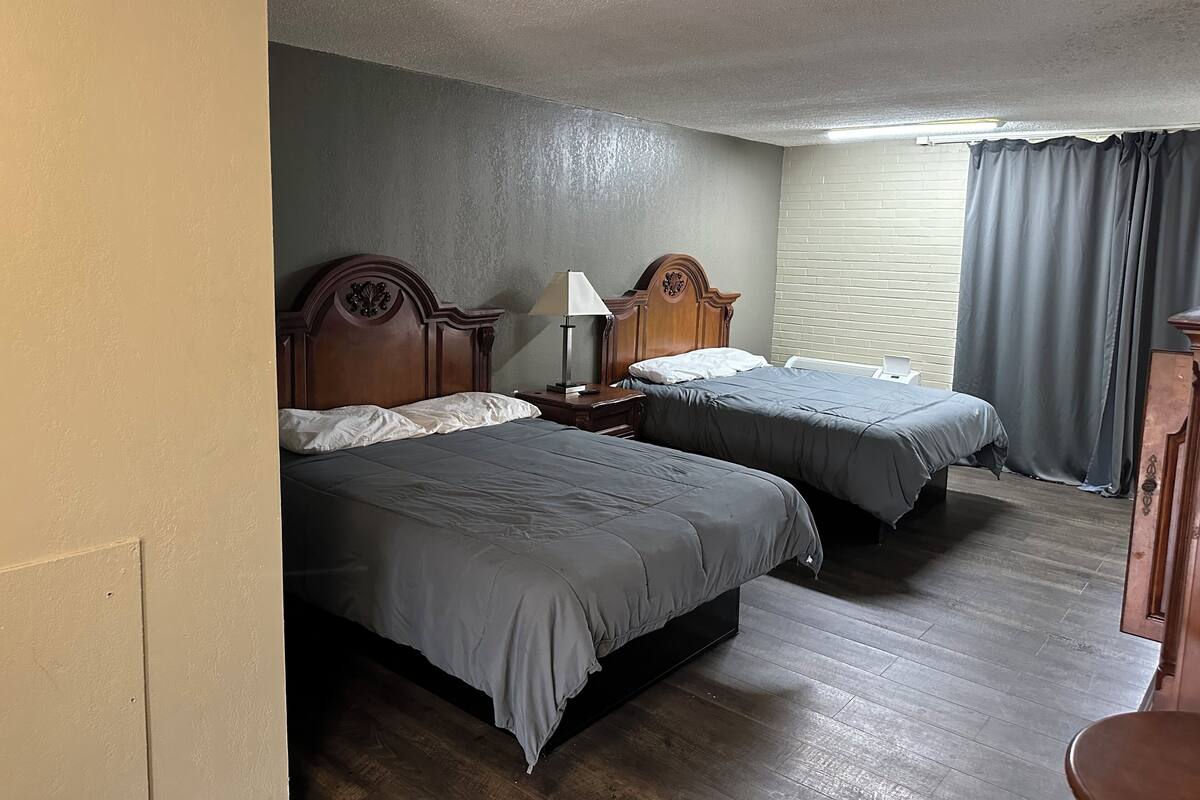 A hotel room with two beds, wooden headboards, a nightstand with a lamp, a window with curtains, and a wooden floor.