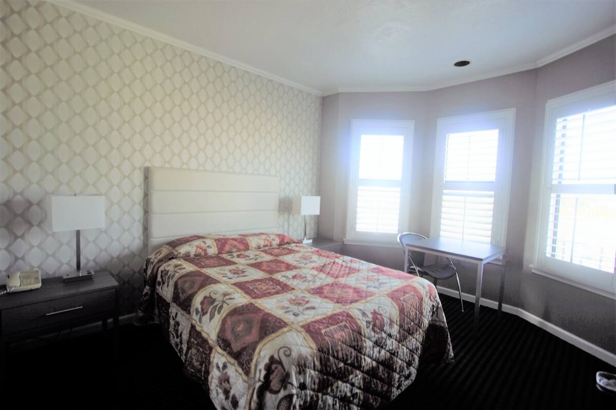 A bedroom with a bed, nightstands, a desk, and two windows.