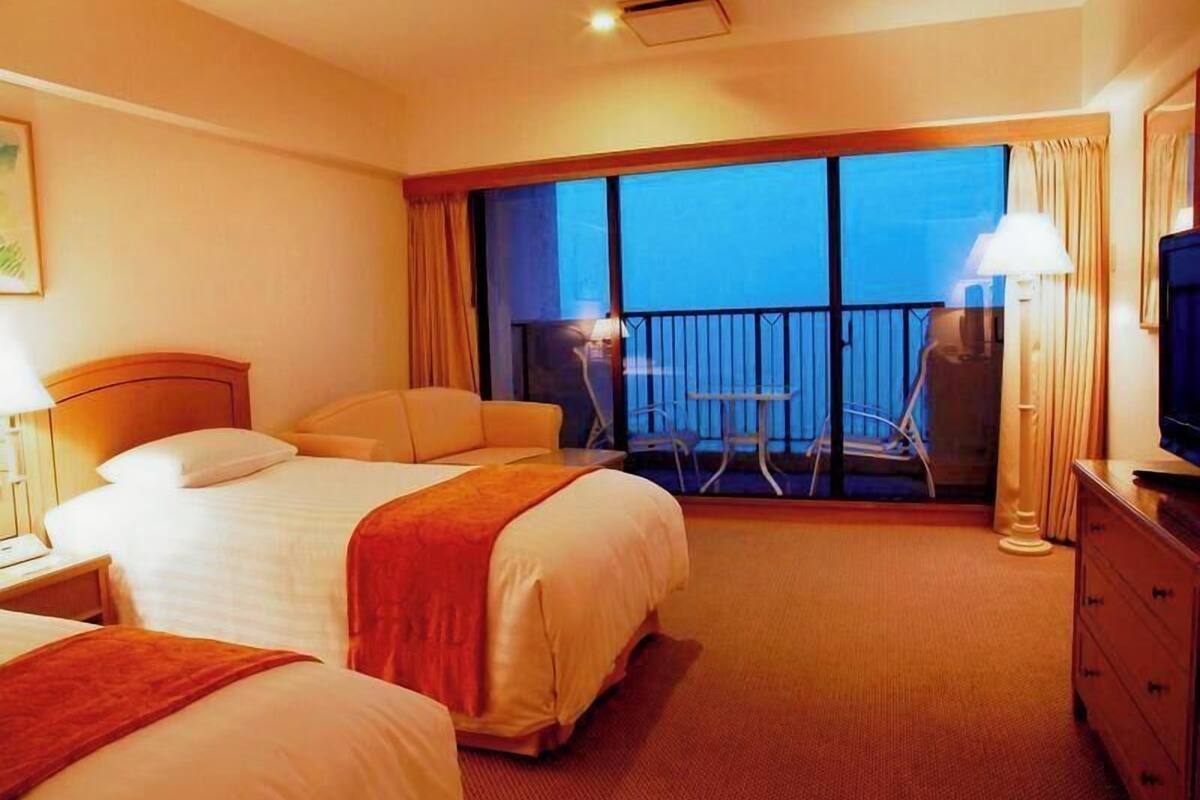 A hotel room with a large bed, a smaller bed, a desk, a chair, a TV, and a balcony with a table and chairs.
